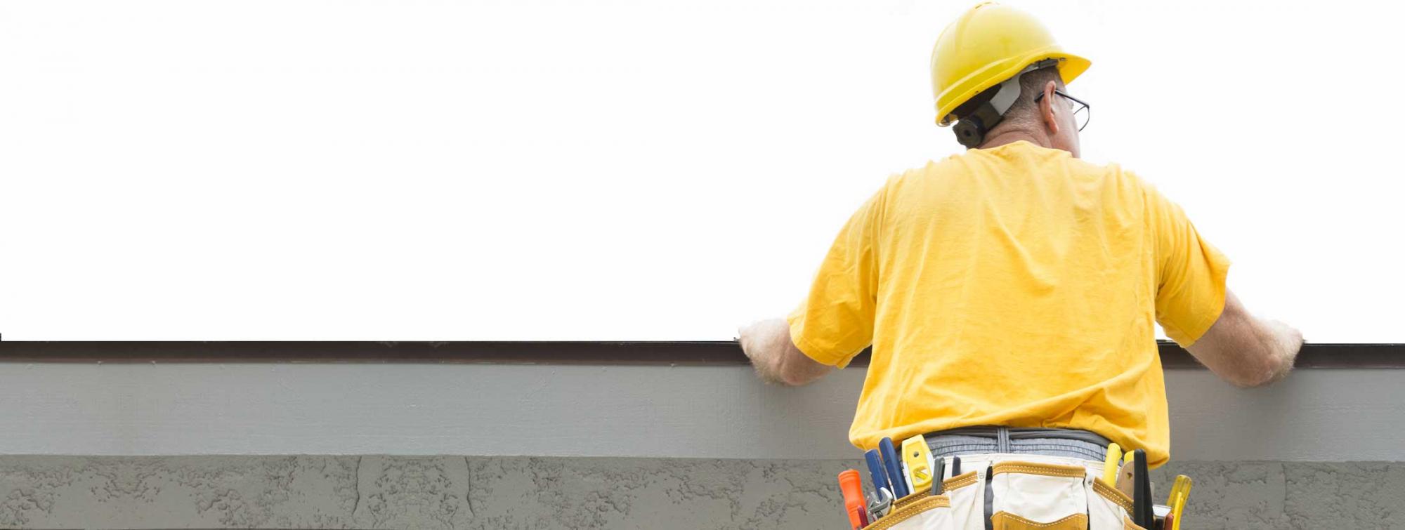 roofer dressed in yellow construction hat and tshirt inspecting a flat roof with tools on his back pocket.