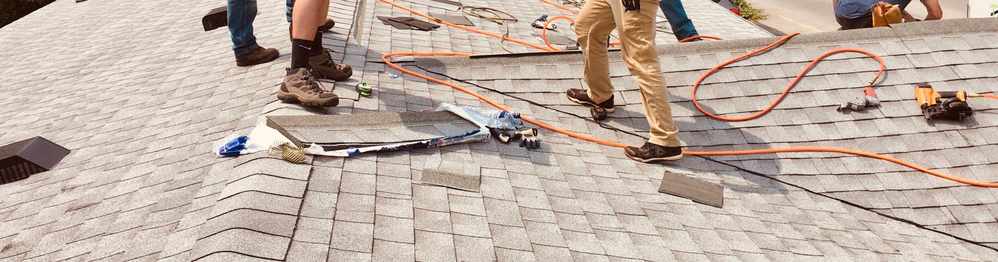roofers repairing grey asphalt shingles being repaired in the afternoon using an electric drill and heavy duty stapler