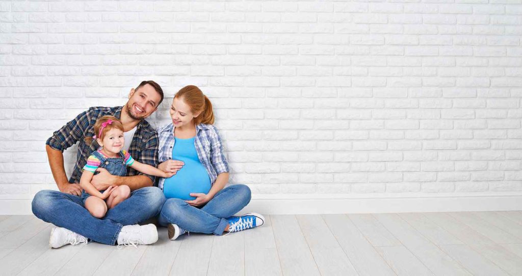 happy family consisting of dad sitting holding his 4 yr old daughter with his pregnant wife beside him seated against white wall