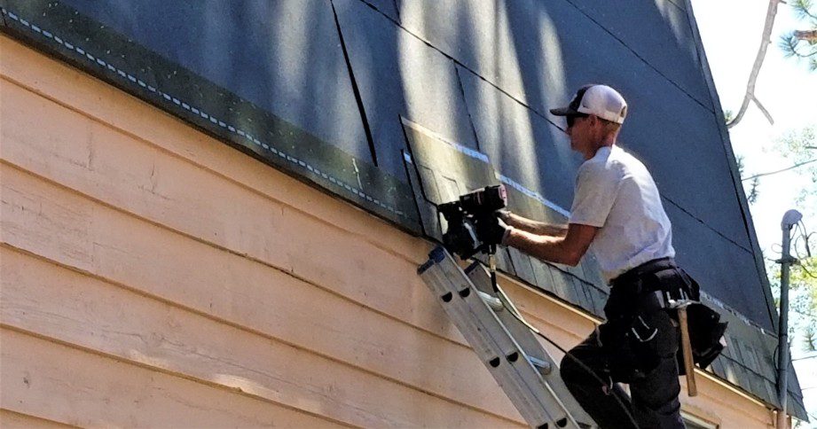 professional roofer on a ladder in black and white uniform replacing rustic black 3-tab asphalt shingles on a roof with a pneumatic nail gun.